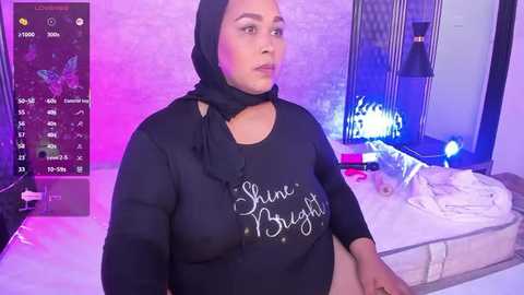 A video of a plus-sized woman with light brown skin, wearing a black hijab and long-sleeved top with \"Shine Bright\" written on it. She sits on a bed in a modern, brightly lit bedroom with purple and blue lighting.