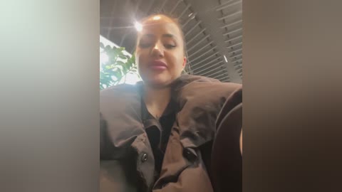 A video of a smiling woman with short, blonde hair, wearing a brown coat, standing in a bright, sunlit room with green plants and horizontal blinds in the background.