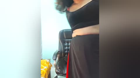 Video of a woman in a black sports bra and high-waisted black pants, seated on a black office chair in a bright room.