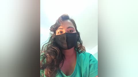 Video of a young woman with long, wavy brown hair, wearing a black face mask and a green shirt, against a blurred white background.