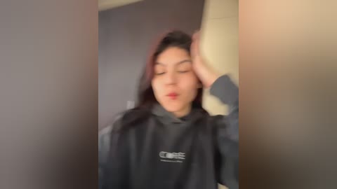 A blurry video of an East Asian woman with long black hair, wearing a dark hoodie, standing in a dimly lit hallway. She has her hand on her head, looking tired.