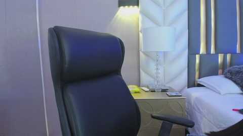 Video of a modern, minimalist hotel room featuring a black leather office chair, a beige nightstand, a white lampshade, a neatly made bed with white sheets, and a dark headboard. The room's decor includes geometric patterns and soft lighting.