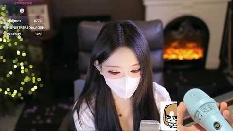 Video of an East Asian woman with long black hair, wearing a white mask, seated in a cozy room with a lit Christmas tree and a fireplace. She's holding a microphone, possibly livestreaming.
