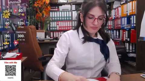 Video of a white woman with long dark hair and red glasses, wearing a white blouse and navy tie, working at a desk in a library filled with colorful books and a potted plant.