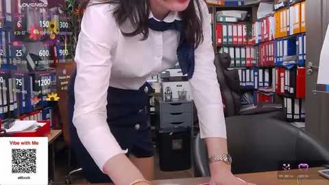 Video of a woman in a white blouse, navy skirt, and blue scarf leaning over a desk in a colorful, cluttered office filled with books, papers, and computer screens.