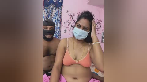 A video of a South Asian woman with a mask, wearing a pink bra, and a man in a black mask and shirt, standing in a pink room with a decorative wall.