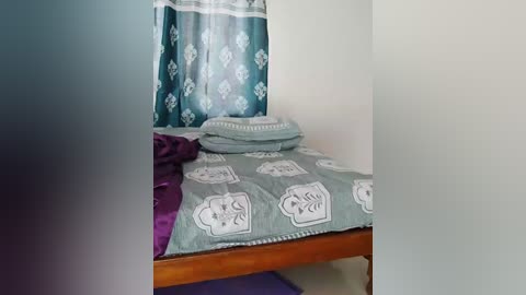 Video of a simple, small bed in a sparsely furnished room. Bed is covered with a green, patterned quilt featuring white geometric designs. Purple blanket is draped over the bed. Background includes a plain white wall and a partially visible teal curtain with floral patterns.