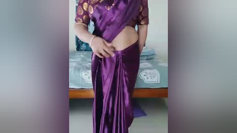 Video of a woman in a purple sari, partially lifting the sari to reveal her bare belly, standing in a simple bedroom with a bed and white curtains in the background.