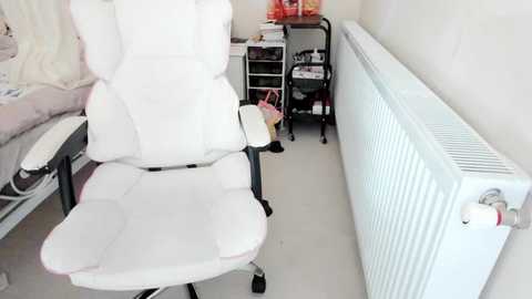 Video of a white, cushioned gaming chair in a minimalist room with a white radiator, black utility cart, and pink storage bags.