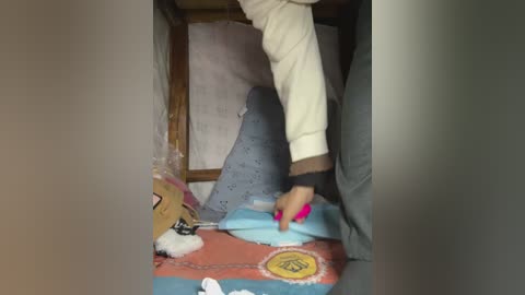 Video of a person, likely a child, dressed in a white sweater, using scissors to cut a blue and orange quilted blanket. The background features a wooden frame with a pink pillow inside.