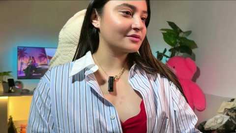 Video of a smiling woman with long black hair, wearing a blue and white striped shirt, a red top, and a black necklace, in a cozy room with a TV, plants, and a pink stuffed toy.