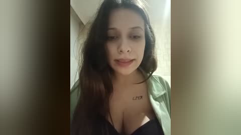 Video of a young woman with long, wavy dark hair, wearing a black bra, green shirt, and a tattoo on her collarbone. She stands against a beige wall, indoors, smiling slightly.