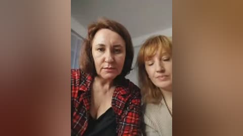 Video of two women in a home setting. The woman on the left, with shoulder-length brown hair, wears a red plaid shirt over a black top. The woman on the right, with blonde hair, wears a beige sweater. Both have neutral expressions.