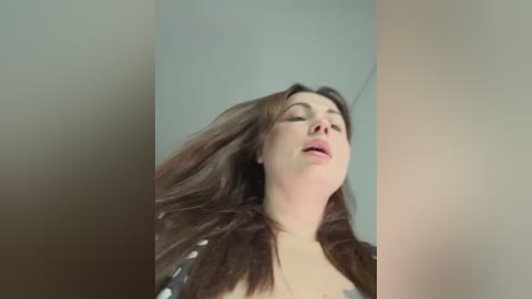 A close-up video of a young woman with long, dark brown hair, fair skin, and closed eyes, wearing a black and white patterned top, captured from a low angle, with blurred background.