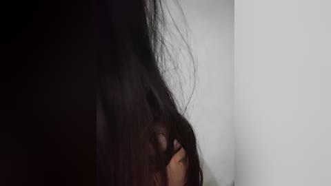 Video of a person with long, dark hair, partially covering their face, standing in a dimly lit room with white walls. The focus is on the hair and the hand gently touching it.