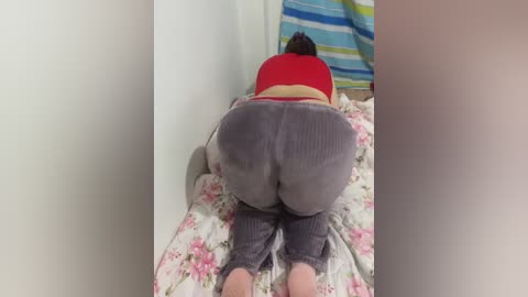 Video of a plus-sized woman with fair skin, wearing a red crop top and grey sweatpants, lying face down on a floral bedspread.