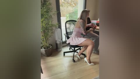 Video of a woman in a pink dress, seated at a desk in a modern office with bamboo plant, wooden floor, and natural light.
