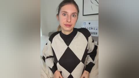Video of a young woman with fair skin, brown hair tied back, wearing a black-and-white diamond-patterned sweater, indoors with minimalist decor and a framed black and white drawing in the background.