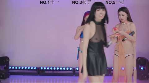 Video of three East Asian women on a stage, wearing revealing, black, see-through dresses, smiling and interacting. Background includes stage lights and Chinese text.