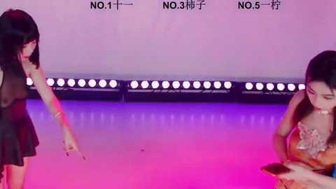 Video of two women in a modern, colorful setting with a pink and purple background. One woman in a black dress is gesturing, and the other in an orange dress is holding a phone.