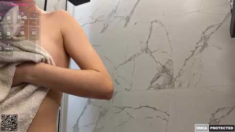 Media: Video of a nude, fair-skinned person with a small to medium-sized breast, holding a white towel in a marble-tiled shower.