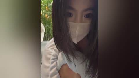 Video of a woman with long, dark hair, wearing a white mask, light blue shirt, and a white top. She is partially obscured by a dark doorway, with green foliage and a white wall visible in the background.