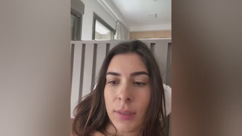 Video of a young woman with medium skin tone and dark brown hair, slightly parted lips, and a neutral expression. She stands in a modern, light-colored bedroom with a window and a white curtain.