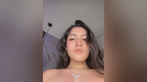 Video of a Latina woman with long, wavy dark hair, light brown skin, and full lips, wearing a diamond necklace. She has a confident, closed-mouth smile. Background is blurred with a hint of a wall.
