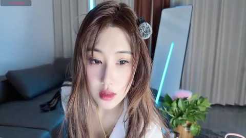 Video of an Asian woman with long brown hair, wearing a white kimono, in a modern living room with a mirror, potted plants, and grey furniture.