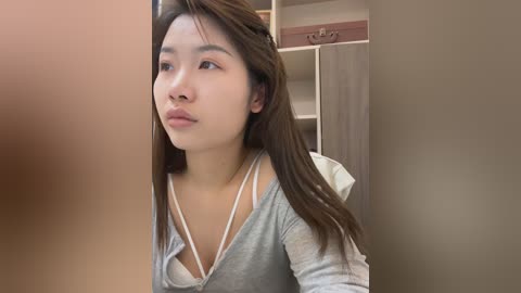 Video of an East Asian woman with long brown hair, wearing a light gray top, looking curious through a door frame. Background includes beige walls and a closet.