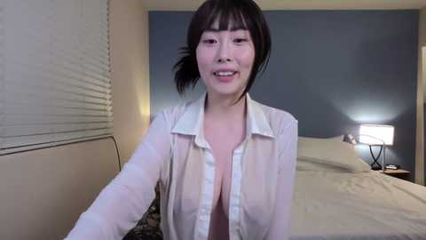 A video of an Asian woman with short black hair, wearing a white, see-through blouse revealing a cleavage, smiling in a dimly lit bedroom with a bed and lamp.