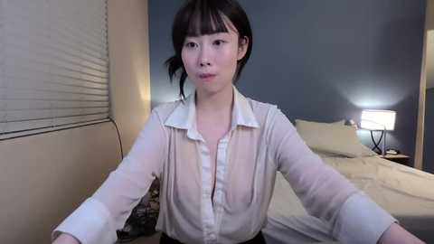 Video of an Asian woman with short black hair, wearing a sheer white blouse, sitting on a bed in a dimly lit room with a blue wall and a bedside lamp.