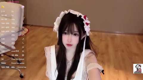 A video of a young East Asian woman in a maid outfit, with a white frilled headpiece, sitting on a wooden floor. The background includes a computer monitor and a wooden chair.