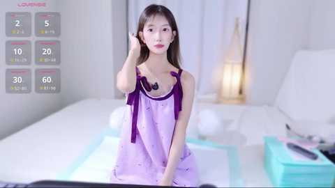 A young Asian woman with long brown hair, wearing a purple nightgown with a ribbon, sits on a white bed in a minimalist bedroom.