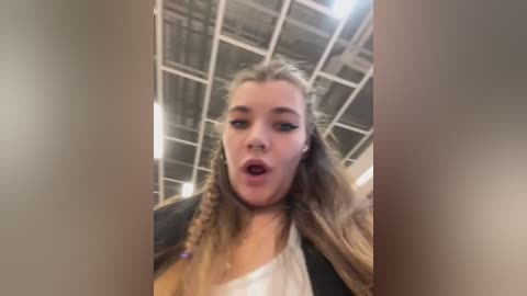 Video of a young woman with long blonde hair in braids, wearing a white tank top, standing indoors with a ceiling made of white panels.