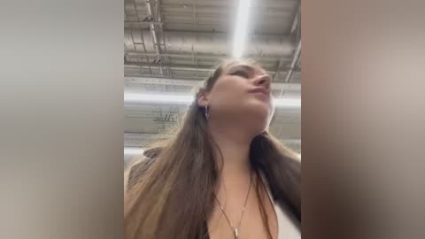 Video of a woman with long, straight brown hair, wearing a brown jacket and a necklace, standing in a warehouse with a high ceiling and exposed pipes.
