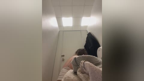 A video of a woman with short brown hair, wearing a gray sweater, sitting on a bed with a white blanket, in a dimly lit, small, white-walled room with a fluorescent ceiling light.