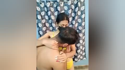 Video of a South Asian woman with dark hair, wearing a yellow sari, kissing a young boy's forehead. She has a mask on. Background shows a patterned shower curtain.