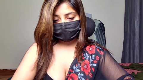Video of a woman with long, straight brown hair, wearing a black floral blouse and a black face mask, sitting on a chair in a dimly lit room with beige walls and a dark curtain.