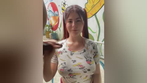 Video of a young woman with long, straight brown hair, wearing a white t-shirt with cartoon characters. Background features colorful, whimsical mural with flowers and insects.