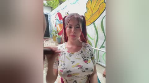 Video of a young woman with fair skin and long, red hair styled in pigtails, wearing a white shirt with cartoon characters, standing in a colorful, graffiti-adorned alleyway.