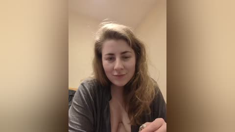 Video of a young, light-skinned woman with wavy brown hair, wearing a grey cardigan, seated in a beige hallway. She has a soft smile and holds a small object in her hand.