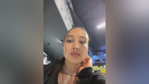 Media: Video of a young woman with medium brown skin, dark hair, and medium-sized breasts, wearing a black jacket, inside a dimly lit room with a blue and white patterned ceiling.