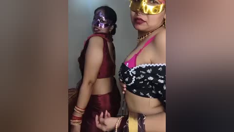 Video of two women in Indian attire; one in a red lehenga and the other in a black choli with a golden mask, standing indoors.