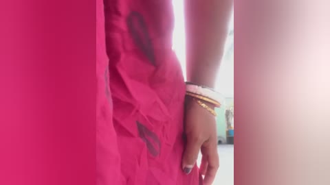 A close-up video of a woman's lower torso in a vibrant pink dress, showing her waist and arm adorned with gold bangles. The background is slightly blurred, with hints of a white wall.