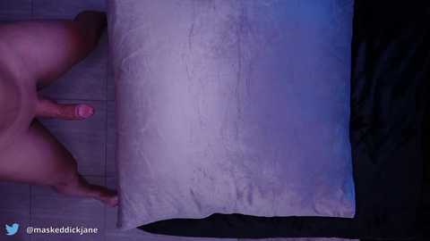A video featuring a close-up of a naked man's erect penis, standing on a wooden floor with a large beige pillow. The background is dimly lit with blue lighting.