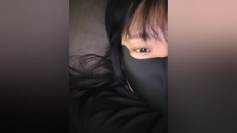 A close-up video of a person with light skin, wearing black clothing, and a black face mask, partially obscuring their eyes, set against a blurred, dark background.