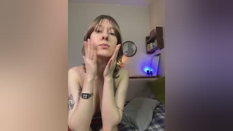 Media: A video of a fair-skinned, small-breasted young woman with short, light brown hair, topless, sitting in a bedroom. She has a tattoo on her left forearm and a Fitbit on her left wrist.