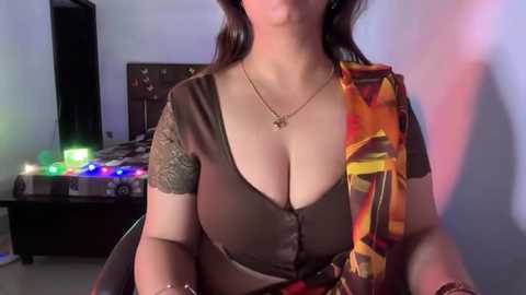 Video of a woman with medium skin tone, wearing a low-cut brown top with a gold chain necklace, sitting in a dimly lit room with colorful string lights and a wooden desk in the background.