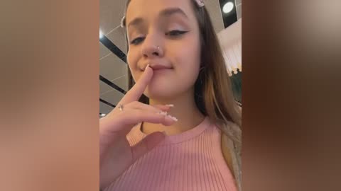 A young woman with light skin, long brown hair, and a nose ring, wearing a pink ribbed top, playfully covers her mouth with her finger. Background shows an indoor, possibly mall setting with modern architecture and soft lighting.